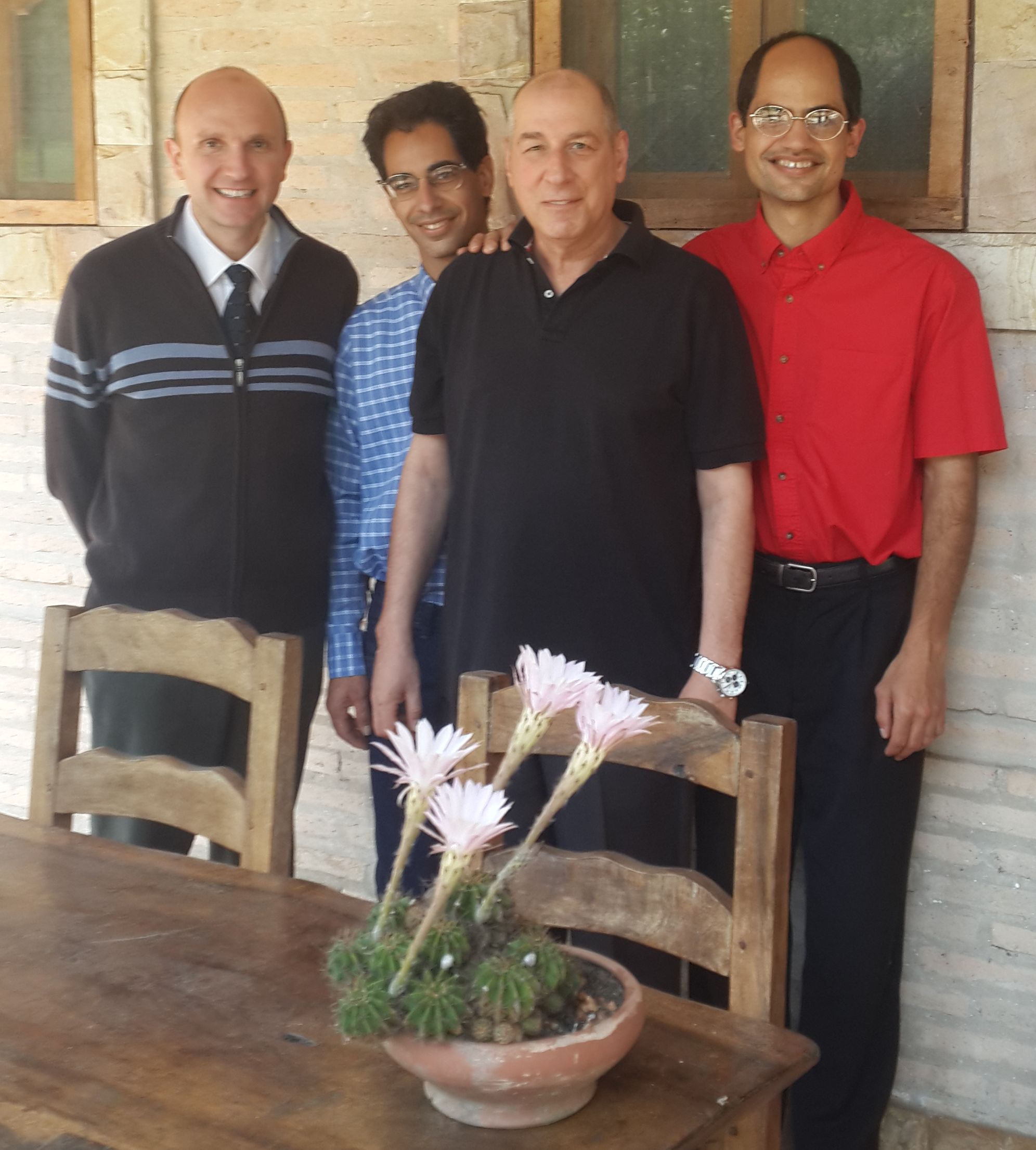 Four men of varying ages, standing together with cheerful expressions in front of a rustic building with wooden window frames and a stone facade. They are casually dressed, some in dark shirts, one in a blue and white striped shirt, and another in a red button-down shirt. In the foreground is a wooden table with a cactus planter displaying several blooming light pink flowers.