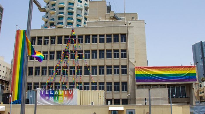 Fotografía de un paisaje urbano con un edificio moderno adornado con múltiples banderas arcoíris y una estructura decorativa que asemeja una pirámide abstracta compuesta por coloridas banderas triangulares. Una pancarta dice &quot;Tel Aviv Celebra el Orgullo&quot;.