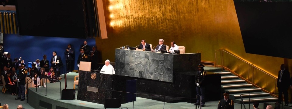 El Papa Francisco se dirige a los delegados en una asamblea de las Naciones Unidas, de pie en un podio en un gran salón con una audiencia y dignatarios sentados y de pie alrededor, debajo de un gran fondo dorado.