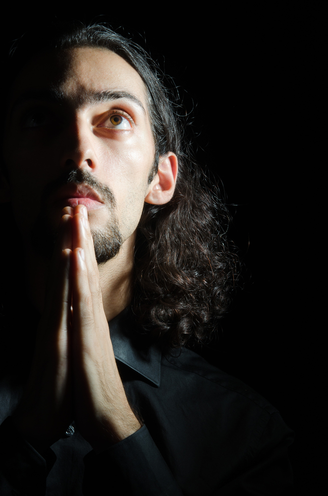 Un hombre de cabello oscuro, largo y rizado, con barba, se representa en actitud contemplativa, con los dedos apretados contra la barbilla en señal de oración. Mira hacia arriba con una mirada concentrada, iluminado por una luz suave y direccional sobre un fondo oscuro.