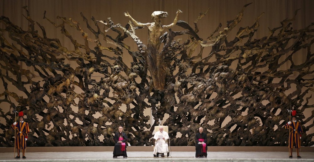 Una gran escultura ornamentada, que asemeja un árbol con extensas ramas, ocupa el fondo. Frente a la escultura se encuentran varias figuras vestidas con diversos atuendos ceremoniales, incluyendo dos con uniformes coloridos que recuerdan a los guardias suizos, dos clérigos sentados, uno de blanco y el otro de morado, y una mujer vestida de negro en silla de ruedas.