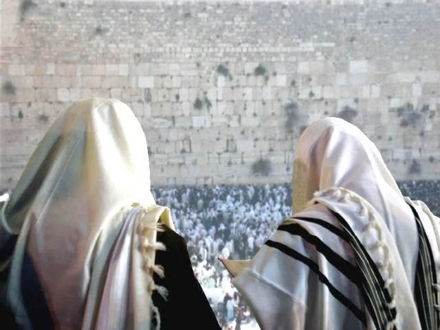 Dos personas con chales de oración judíos tradicionales, de espaldas a la cámara, observan una gran reunión de personas en el Muro Occidental de Jerusalén.