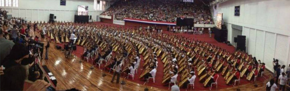 A large indoor graduation ceremony with graduates seated in rows, wearing traditional gowns and caps. The audience fills the surrounding stands, and photographers are capturing the event.
