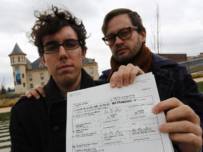 Dos hombres, uno con gafas y el otro con barba, sostienen un documento frente a la cámara, con un edificio y un día nublado de fondo.