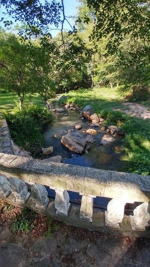 Ein ruhiger Bach fließt durch einen üppigen Park, der von einem rustikalen Steingeländer begrenzt wird. Sonnenlicht fällt durch das dichte Blätterdach und beleuchtet Felsen und Wellenmuster auf der Wasseroberfläche.