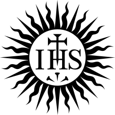 Monochromes Emblem mit den Buchstaben „IHS“ in stilisierter Schrift auf einem kreisförmigen weißen Hintergrund, umgeben von Schwarz. Unter den Buchstaben befindet sich eine dreizackige Form, die an die abstrakte Darstellung einer Krippe oder einer einfachen Krone erinnert.