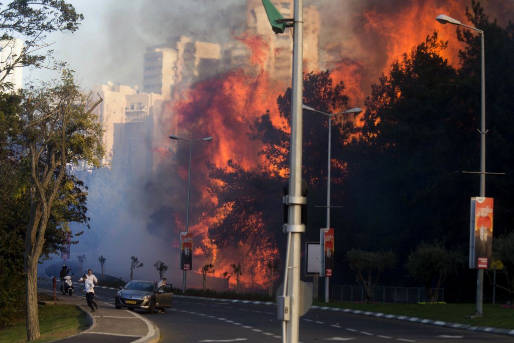 Eine dramatische Szene in der Stadt zeigt ein großes Feuer, das Bäume und Gebäude an einer viel befahrenen Straße verschlingt. Rauch steigt auf, während Fußgänger und Fahrzeuge in der Nähe von Straßenlaternen und Schildern reagieren.